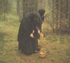 Older women wearing black dresses and head coverings with a basket on the ground in the woods